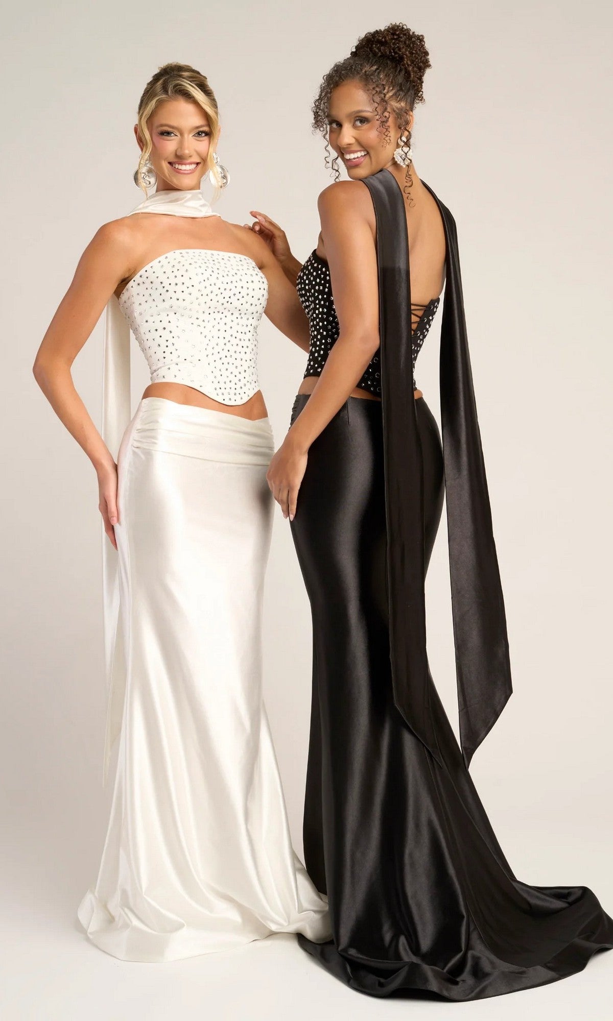 Two women pose in Portia and Scarlett PS26042 long formal dresses: one wears a white beaded corset top with satin skirt, the other a black strapless top and skirt; both embellished with back sashes.